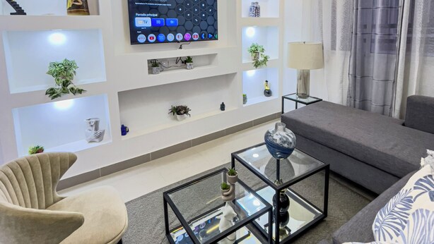 Een moderne woonkamer met een flatscreen televisie aan de muur, een glazen salontafel, een bank en decoratieve items op planken.