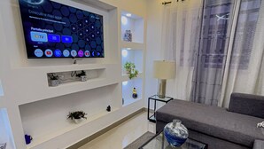 Een moderne woonkamer met een flatscreen televisie aan de muur, een glazen salontafel met decoratieve objecten, een grijze bank en een lamp.