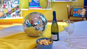 A room with a disco ball, a bottle of champagne, and two glasses on a table. A bookshelf with books and a TV displaying the Netflix logo is in the background. - La Belle Breisach - PopArty - Colmar/Germany (Volgelsheim)