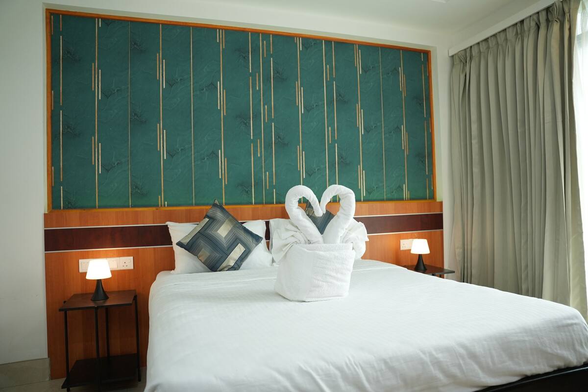 A hotel room with a large bed, a headboard with vertical lines, two bedside tables with lamps, and a large window with curtains.