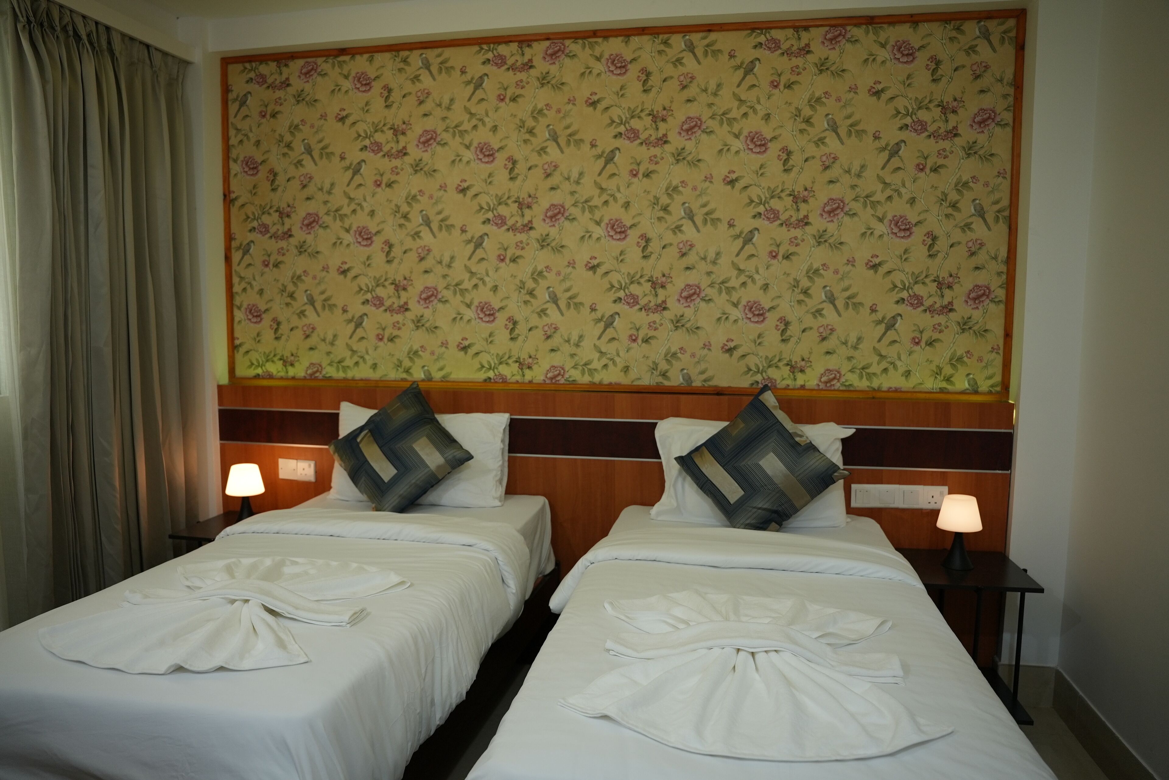 Une chambre d’hôtel avec deux lits, chacun agrémenté d’une serviette blanche soigneusement pliée, d’une tête de lit en bois et d’un papier peint à motifs floraux.