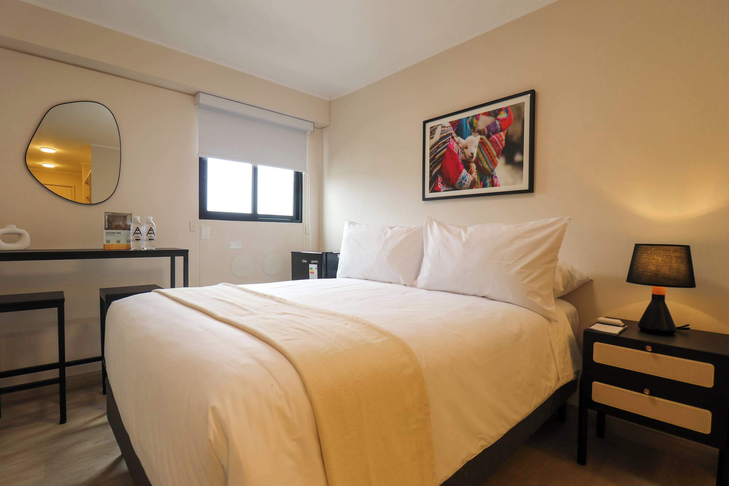 Quarto de hotel com cama, mesa de cabeceira, luminária, escrivaninha, cadeira e um espelho grande.
