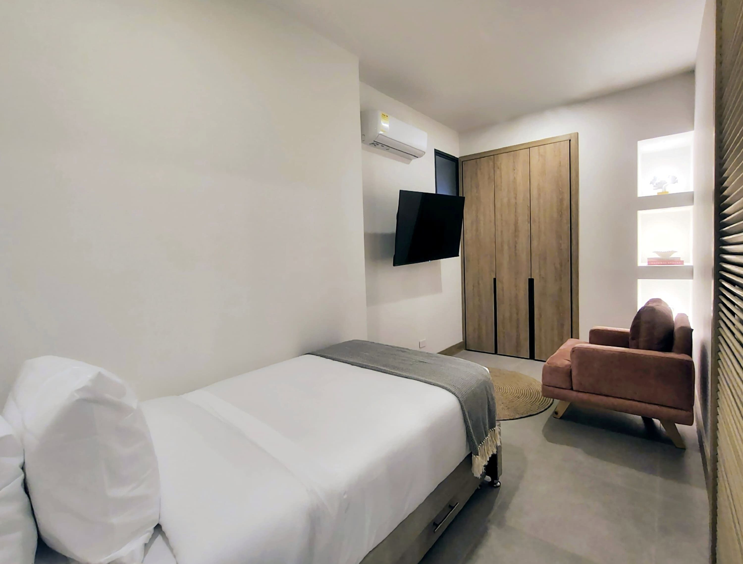 A modern bedroom with a single bed, a TV mounted on the wall, a wardrobe, and a chair.