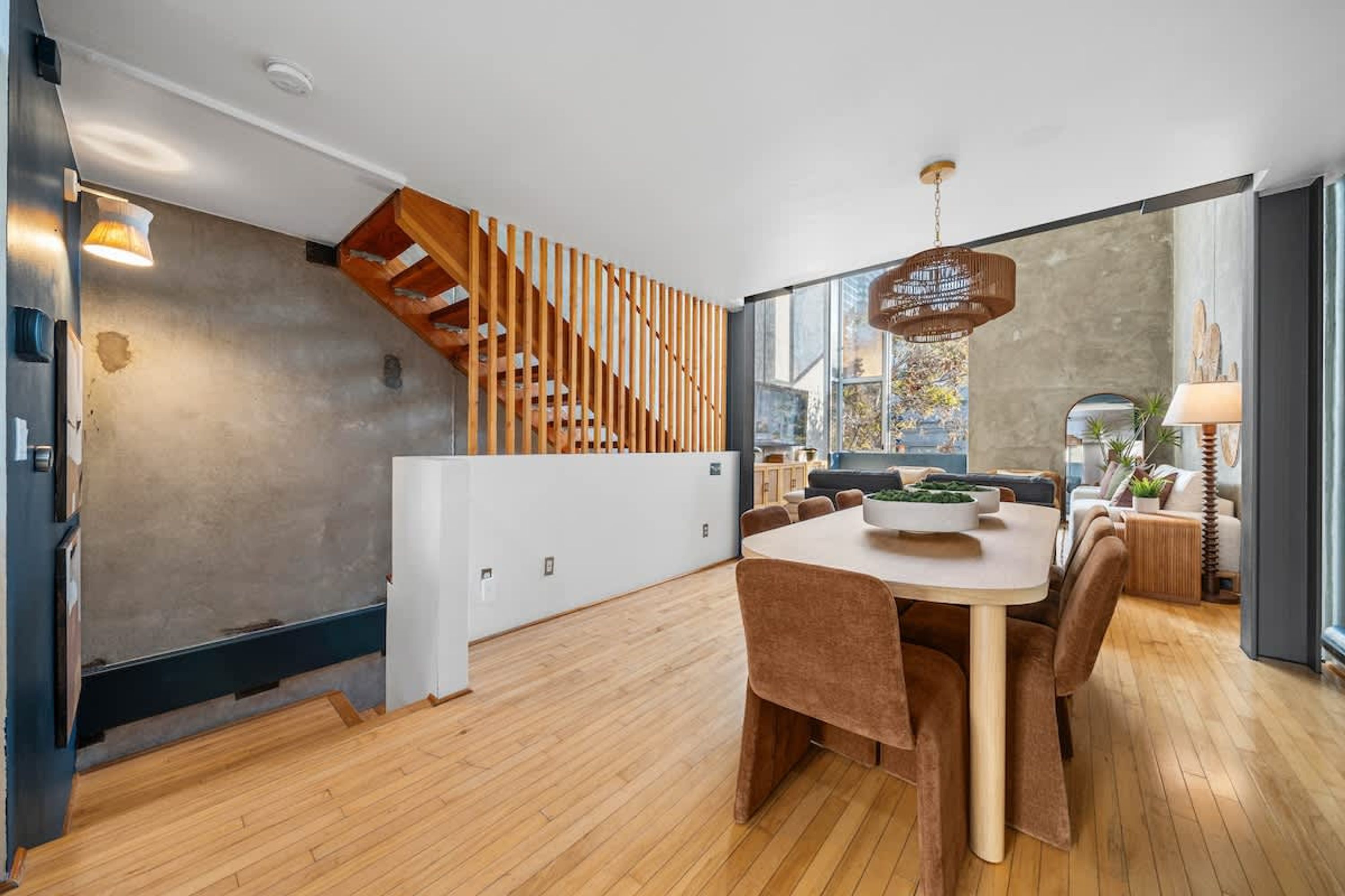 A modern kitchen with a wooden staircase, a dining area with brown chairs, and a large window with a city view.