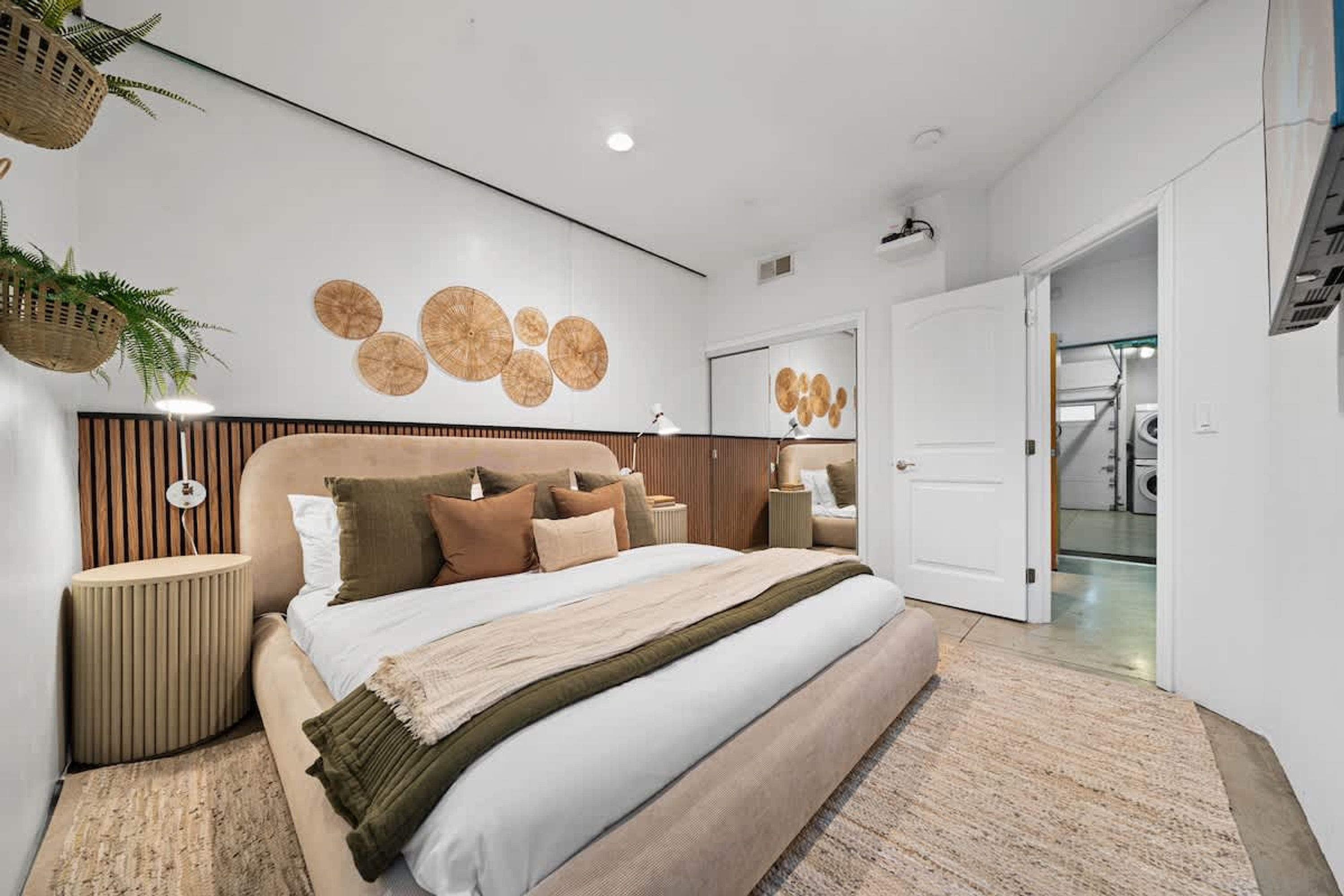 A modern bedroom with a large bed, wooden headboard, bedside tables, and a wall-mounted mirror.
