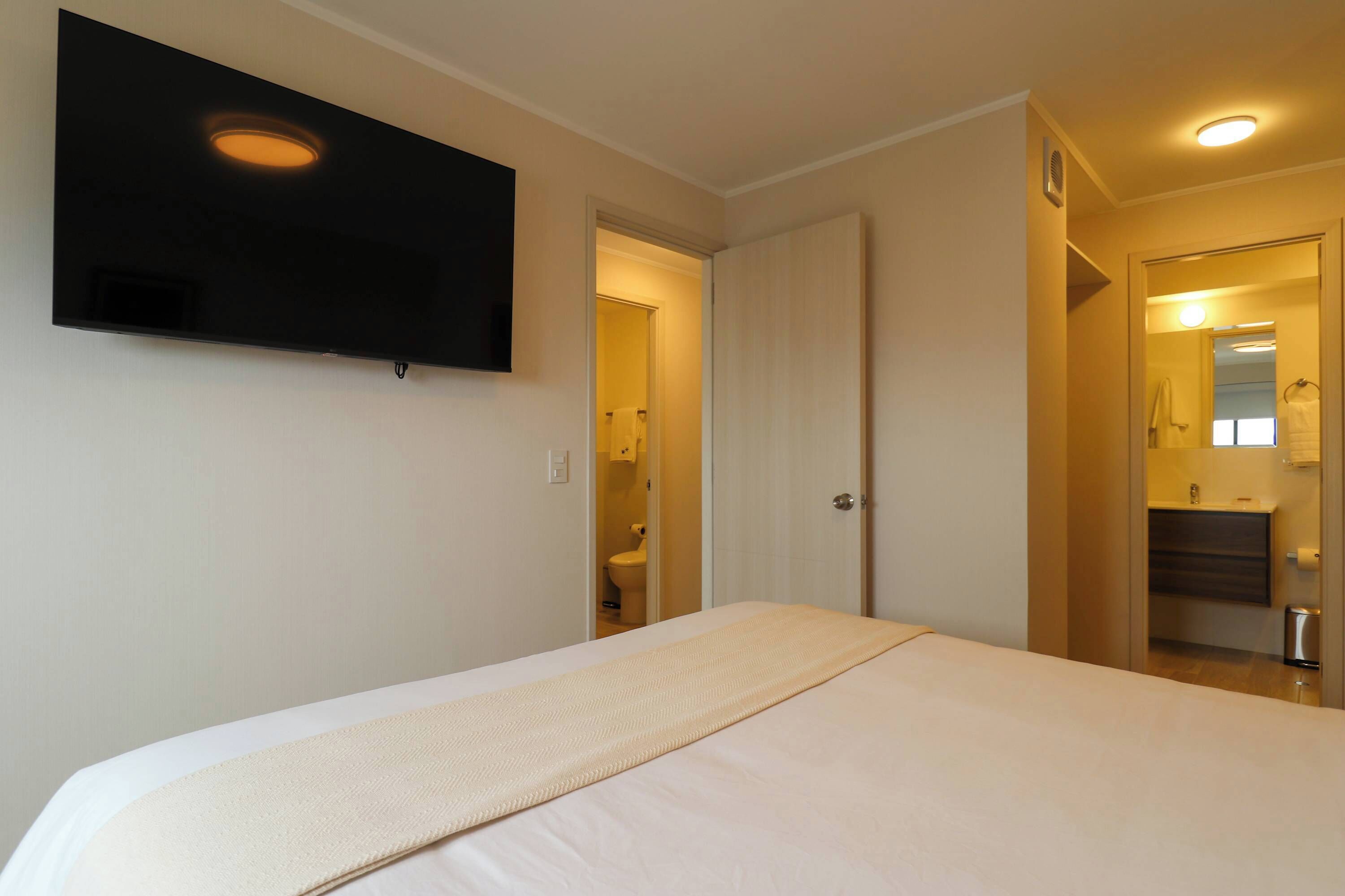 A hotel room with a bed, a TV mounted on the wall, and a bathroom visible through an open door.