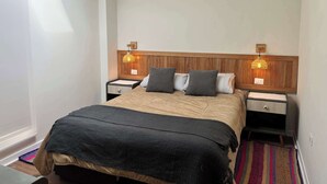 Habitación de hotel con cabecera de madera, dos lámparas de noche, una cama con manta oscura y una alfombra colorida.