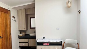 Une chambre avec une porte en bois, un lit, une commode, une chaise, un luminaire suspendu et une petite salle de bain avec un lavabo et un miroir.