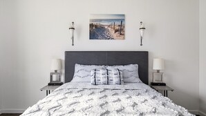Un dormitorio con una cama grande, cabecera, dos mesitas de noche con lámparas y un cuadro enmarcado en la pared.