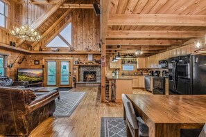 Un interior de cabaña espacioso con techo de madera, chimenea, zona de cocina y sala de estar con sofá de cuero y televisor.