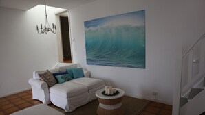 Un salon avec un canapé blanc, un pouf blanc, une petite table d’appoint en osier ornée de bougies, et un grand tableau représentant une vague accroché au mur.