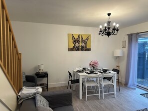 A modern living room with a dining area, featuring a staircase, a chandelier, a dining table set for four, and a large window with a view. - Bright Modern Entire Open-Plan Home –with off street Parking Included (Exeter)