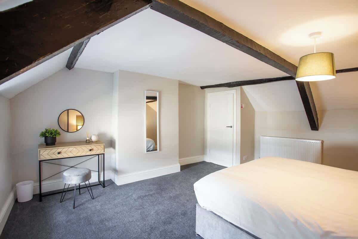 Een slaapkamer met een bed, een kaptafel met spiegel, een klein krukje, een radiator en een plafond met zichtbare balken.