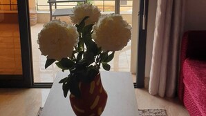A room with a wooden table and a vase of flowers. There is a view of a balcony with a railing and greenery outside.