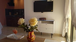 A hotel room with a TV mounted on the wall, a small desk with a decorative item, a vase with flowers, and a glass of orange drink on a tray.