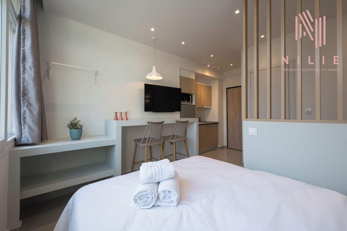 Moderni hotellihuone, jossa on sänky, työpöytä kahdella tuolilla, taulutelevisio ja pieni keittiönurkkaus.