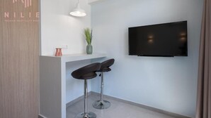 Una zona de recepción moderna de hotel, con una televisión de pantalla plana montada en la pared, dos taburetes de bar, una lámpara colgante y una pequeña planta en maceta.