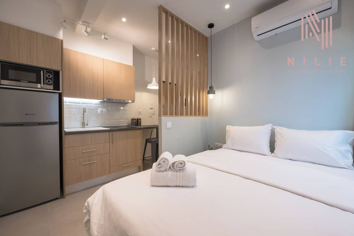Moderni hotellihuone, jossa on pieni keittiönurkkaus, valkoisilla lakanoilla varustettu sänky ja puinen sermi.