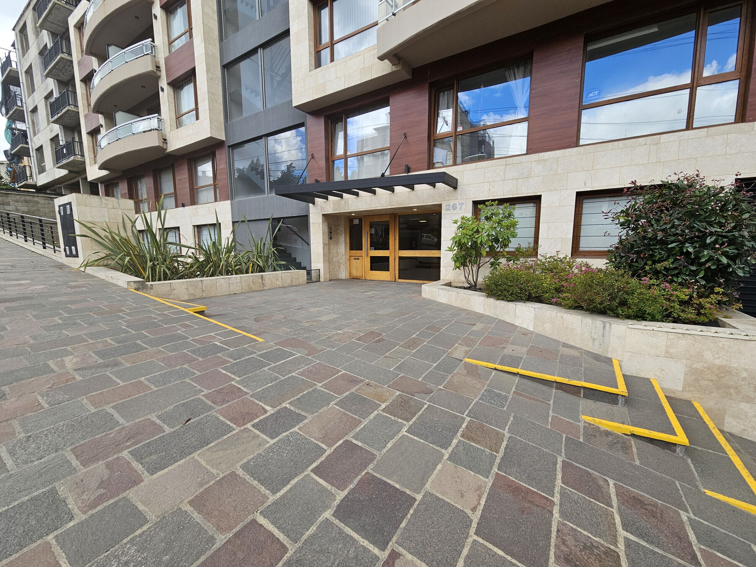 A multi-story building with a modern design, featuring a ground-level entrance with a glass door and a small garden area with plants.