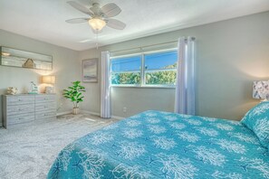 Une chambre avec un grand lit, un ventilateur de plafond, une fenêtre avec des rideaux, une commode et une plante.
