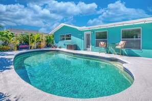 Une piscine à bord incurvé, une maison aux murs extérieurs de couleur turquoise et un espace patio avec des chaises et une table.