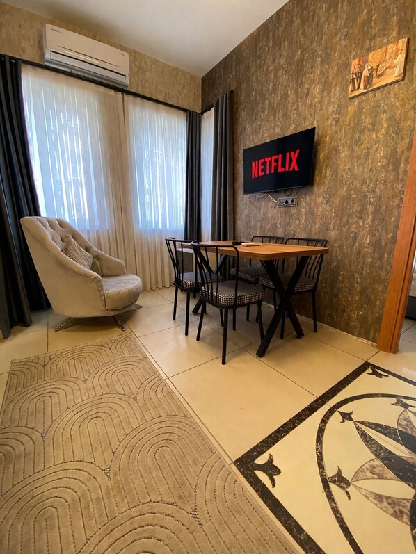 Un salon moderne avec un fauteuil beige, une table à manger en bois avec des chaises, un téléviseur à écran plat fixé au mur et un tapis à motifs.