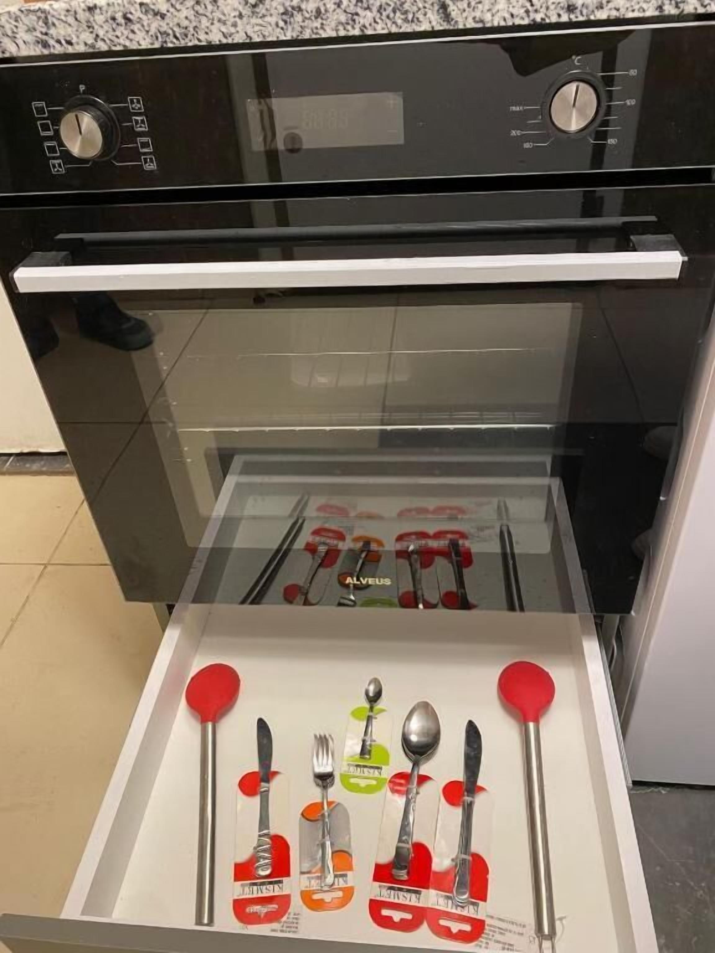 An open oven with a drawer below containing neatly arranged cutlery sets.