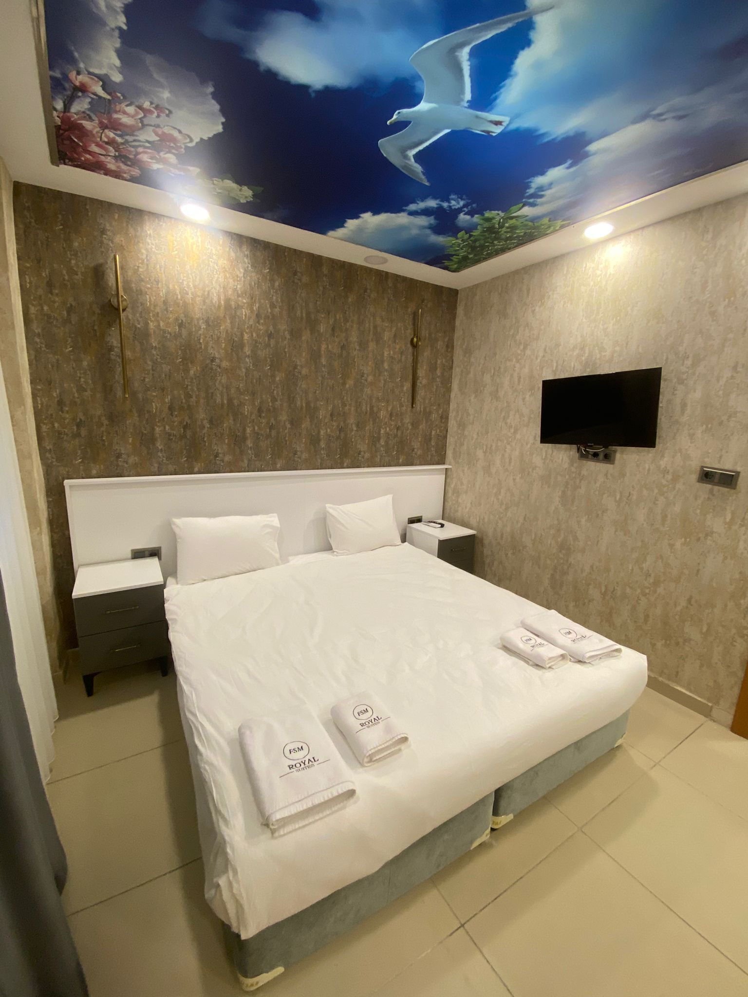 A modern hotel room with a large bed, white linens, a flat-screen TV mounted on the wall, and a mural of a seagull flying above a floral sky.
