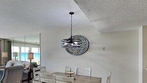 Un coin repas avec une table en bois et des chaises blanches, un élément décoratif mural et une vue sur l’océan par de grandes fenêtres.