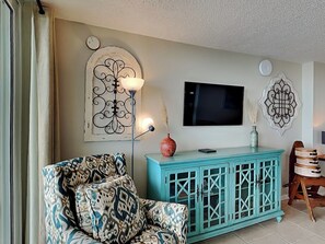 A living room with a patterned armchair, a teal cabinet, a flat-screen TV, decorative wall art, and a small table with a vase. - Dunes of Panama #B301 | Gulf Front + Amenities (Panama City Beach)