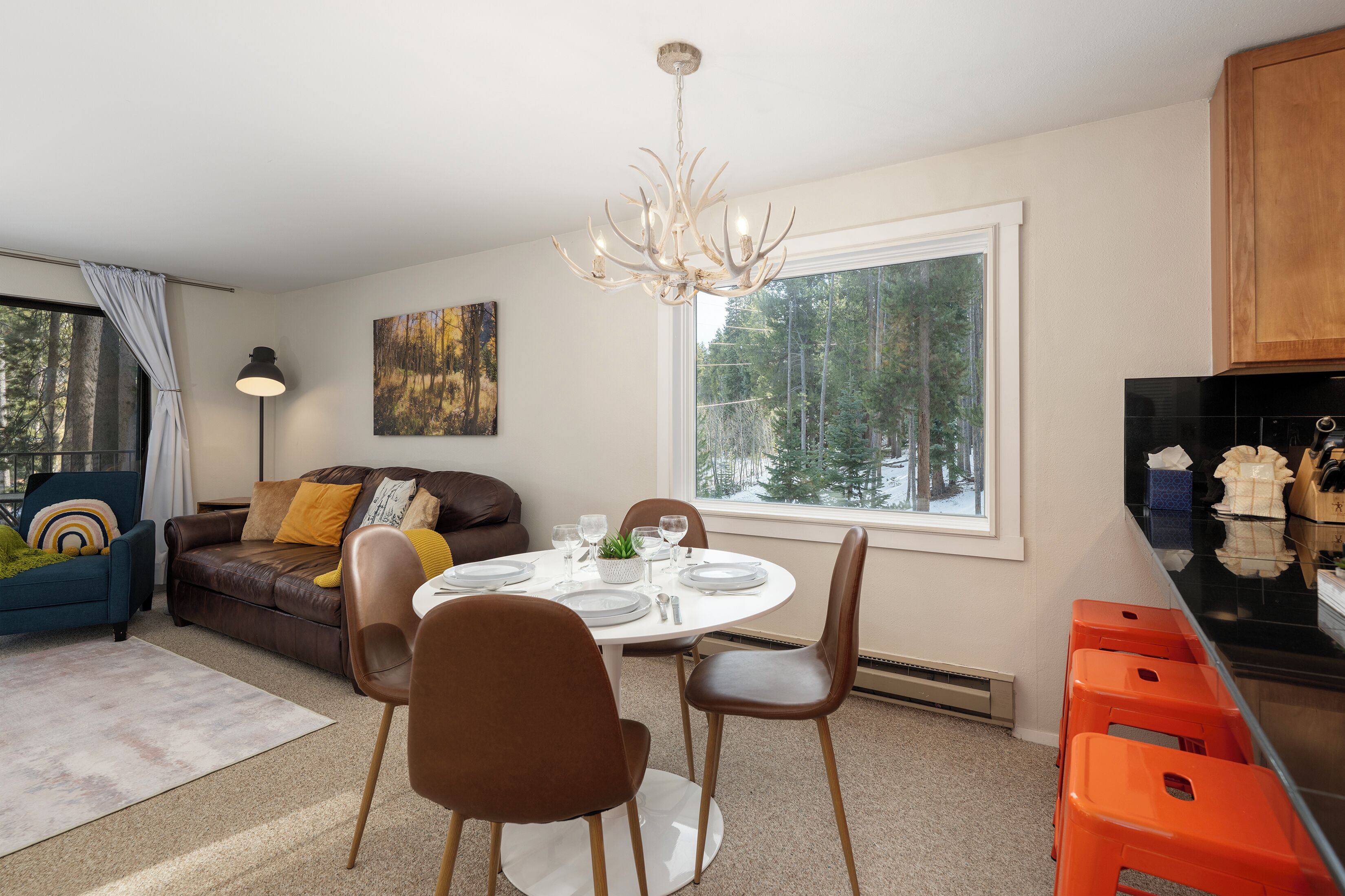 Een moderne woonkamer met een eetgedeelte, een groot raam met uitzicht op bomen, en een kroonluchter.