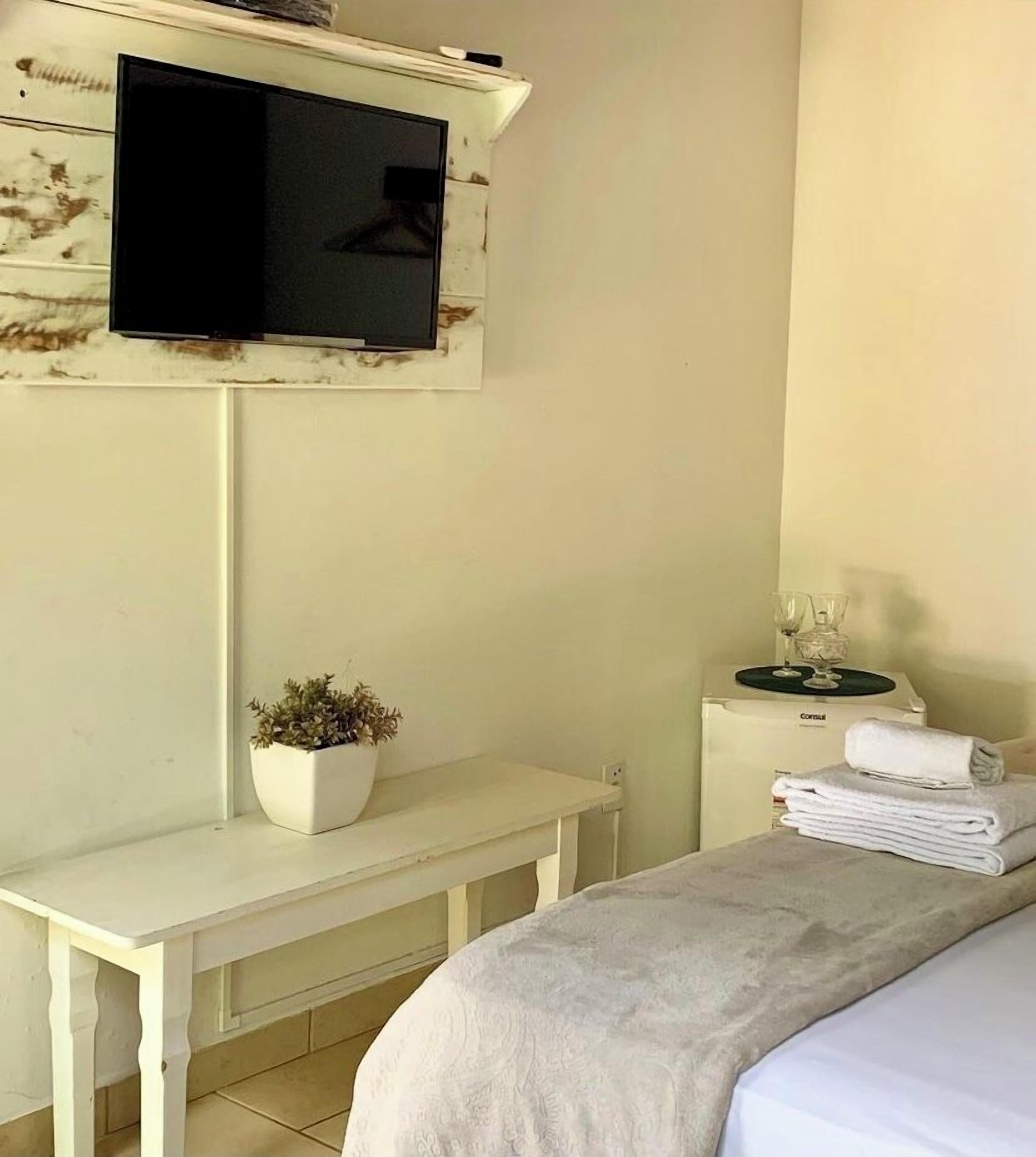A bedroom with a bed, a small desk with a plant, a TV mounted on the wall, and a bedside table with a glass and a towel.