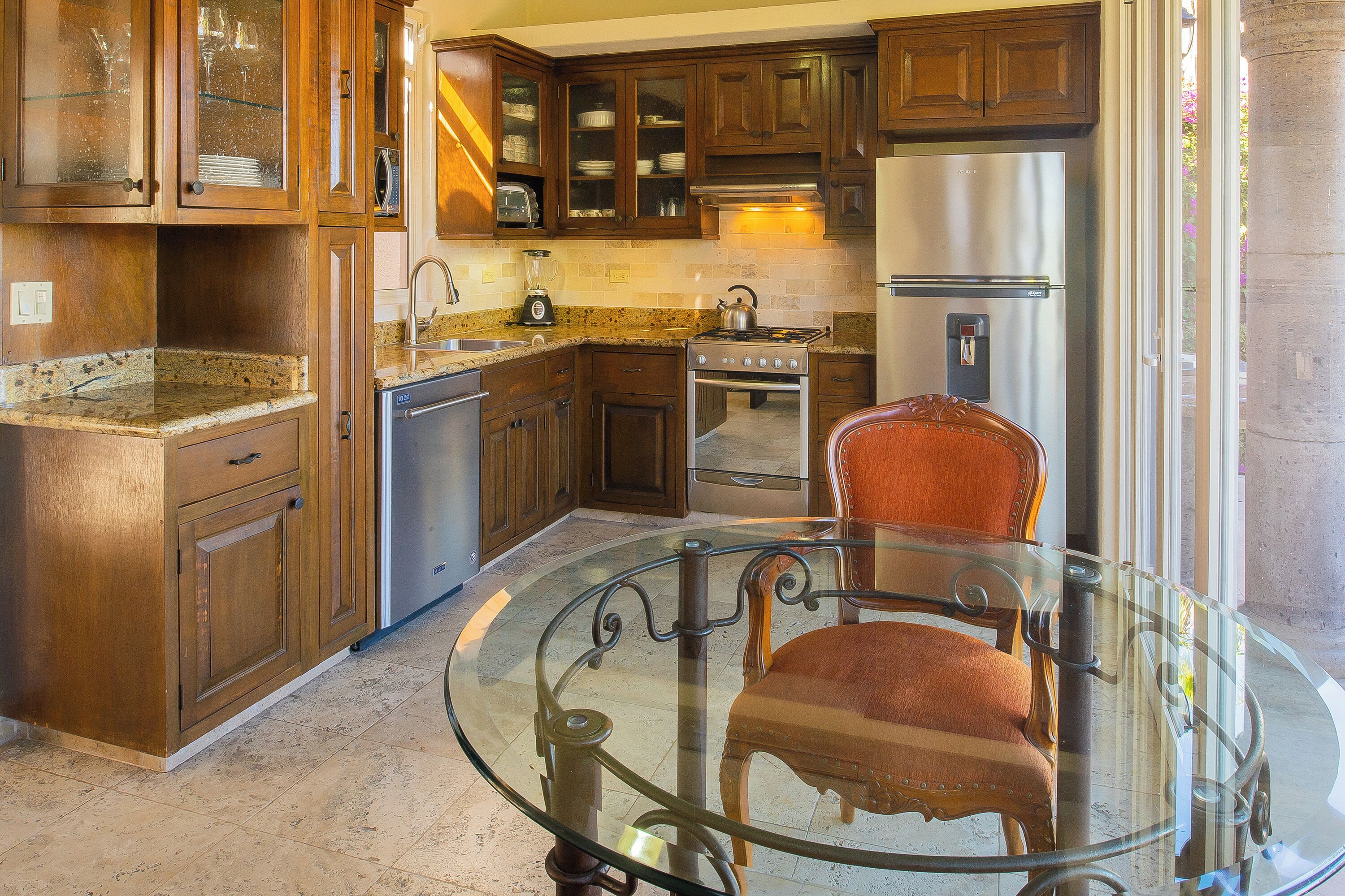 A kitchen with wooden cabinets, a stainless steel refrigerator, a stove, and a glass table with two chairs.