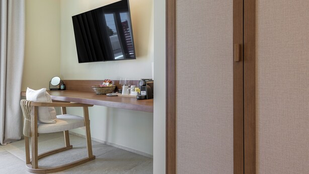 A modern hotel room with a wooden desk, a chair, a flat-screen TV mounted on the wall, and a wardrobe. - HOTEL ELENI (Milos)