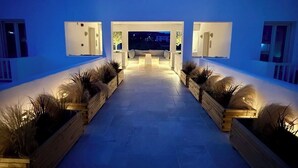 A modern outdoor lounge area with a central seating arrangement, flanked by wooden planters with tall grasses, and illuminated by ambient lighting. - HOTEL ELENI (Milos)