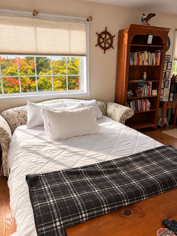 A bedroom with a large bed, a window with blinds, a bookshelf, and a wall-mounted ship's wheel.
