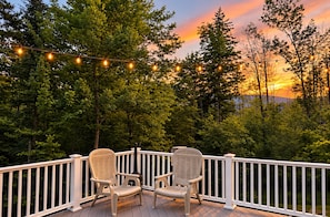 A deck with two wicker chairs and a string of lights against a backdrop of trees and a sunset.