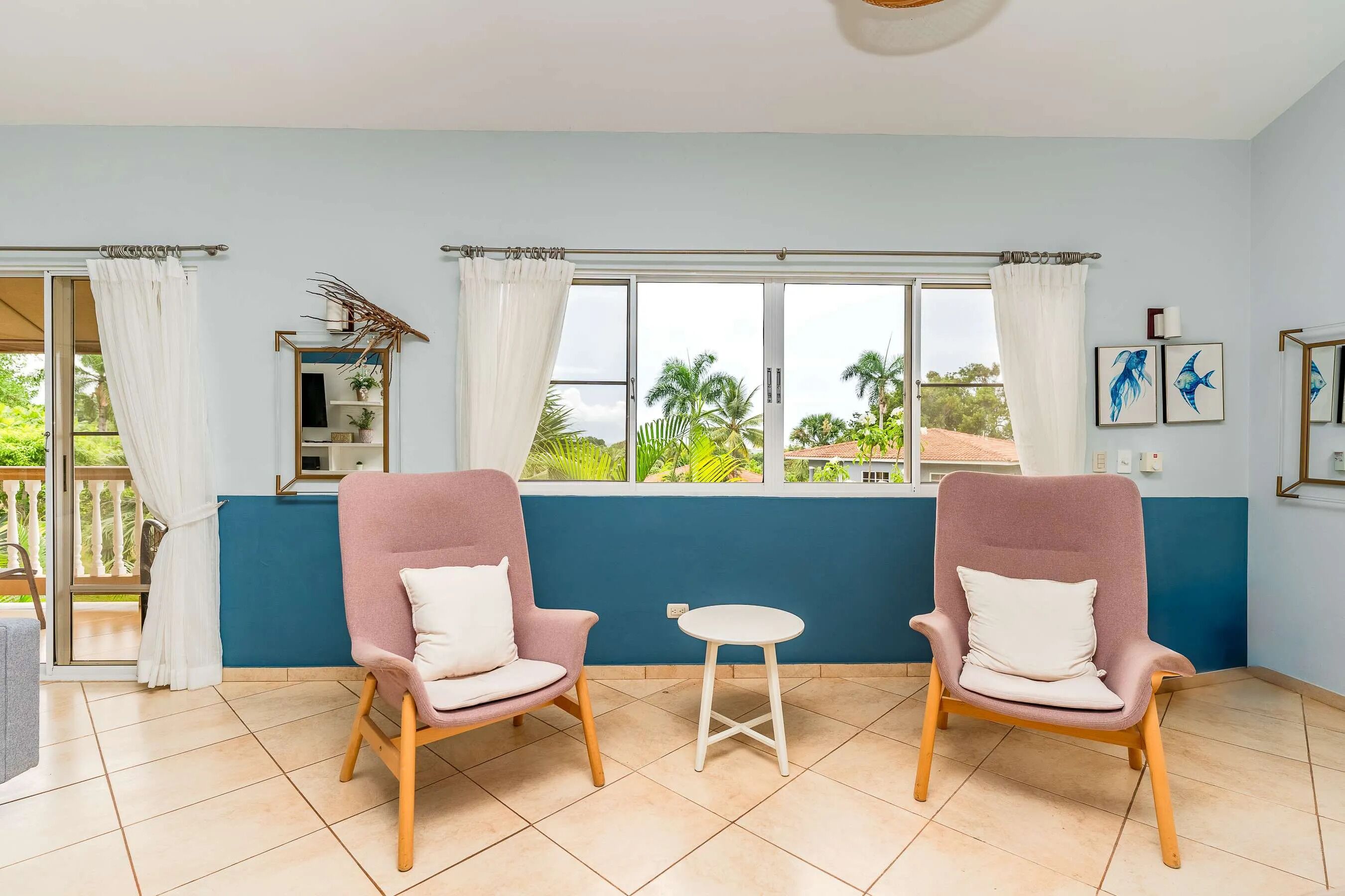 A room with two pink armchairs, a small white table, and a view of greenery through large windows.
