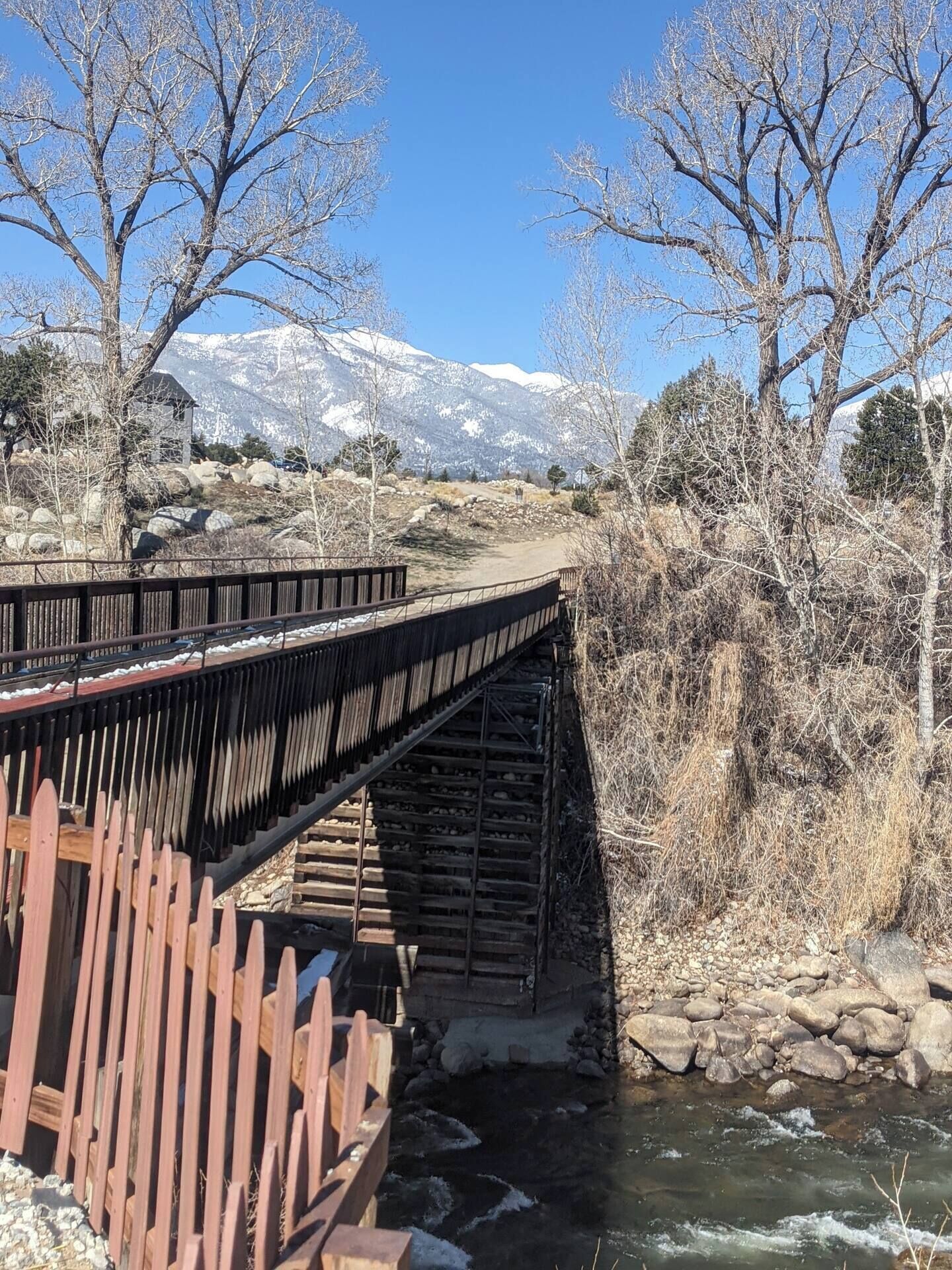 Un pont de fusta sobre un riu, amb una riba rocosa i muntanyes nevades al fons.
