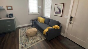 A living room with a dark grey sofa, yellow cushions, a patterned rug, a coffee maker on a counter, and a framed picture on the wall.