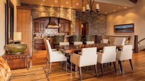 Un comedor espacioso con una gran mesa de madera y sillas blancas tapizadas, un muro de acento de piedra y una cocina moderna al fondo.
