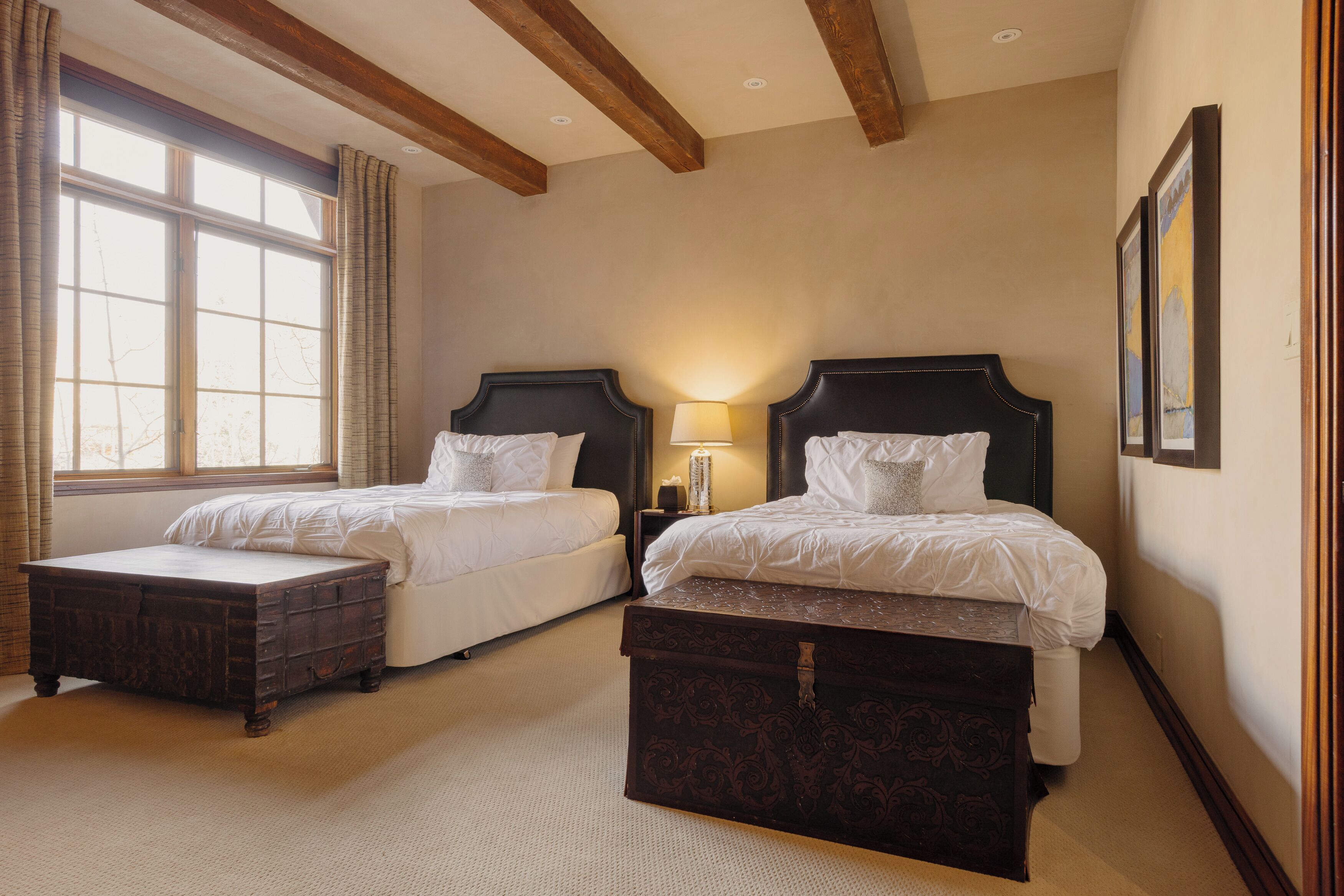 Quarto de hotel com duas camas, uma janela ampla, vigas de madeira e uma mesa de cabeceira com abajur.