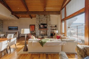 A spacious living room with a stone fireplace, wooden ceiling beams, and large windows offering a city view. - See Forever Penthouse by InvitedHome | Ski Valet, Walk to Ski, Views, Fireplace (Mountain Village)
