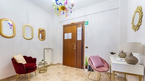 Un hall d’hôtel avec un fauteuil rouge, un fauteuil rose, un bureau blanc avec des lampes et une porte en bois portant un panneau indiquant la sortie.