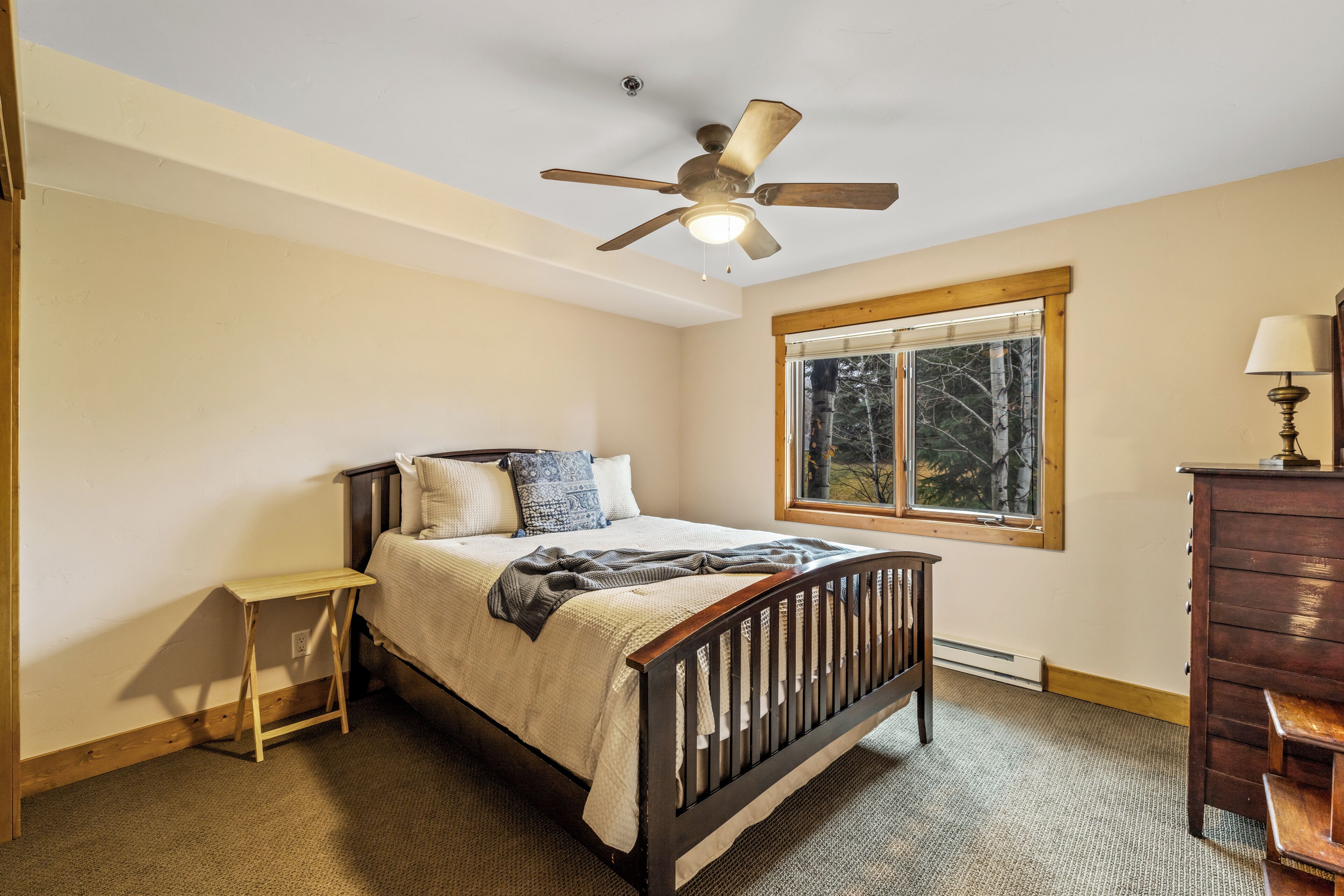 A bedroom with a wooden bed, a nightstand, a dresser, a ceiling fan, a window with a view of trees, and a lamp.