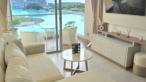Una sala de estar moderna con un sofá grande, una mesa redonda y un televisor de pantalla plana. La habitación tiene ventanas amplias que ofrecen vistas a una piscina y edificios de un complejo turístico.