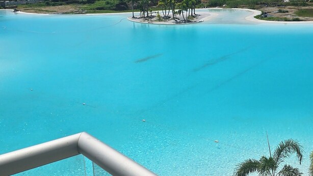 Vista desde un balcón sobre una piscina azul cristalina, una playa de arena y palmeras.