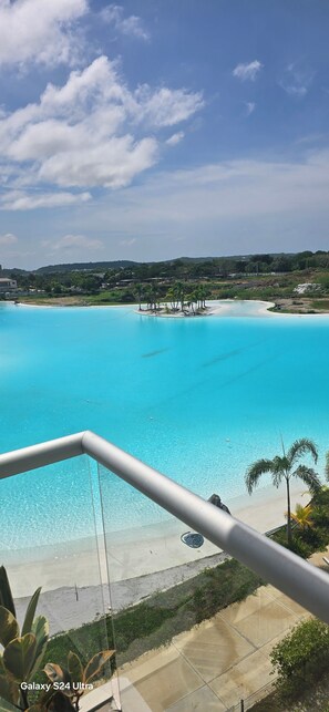 Une vue depuis un balcon donnant sur une piscine d’eau cristalline, une plage de sable et des palmiers.