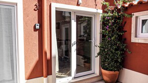 Puerta corrediza de vidrio que da a un cuarto con vista a la calle, una planta en maceta con flores moradas y un dispositivo de seguridad en la pared.
