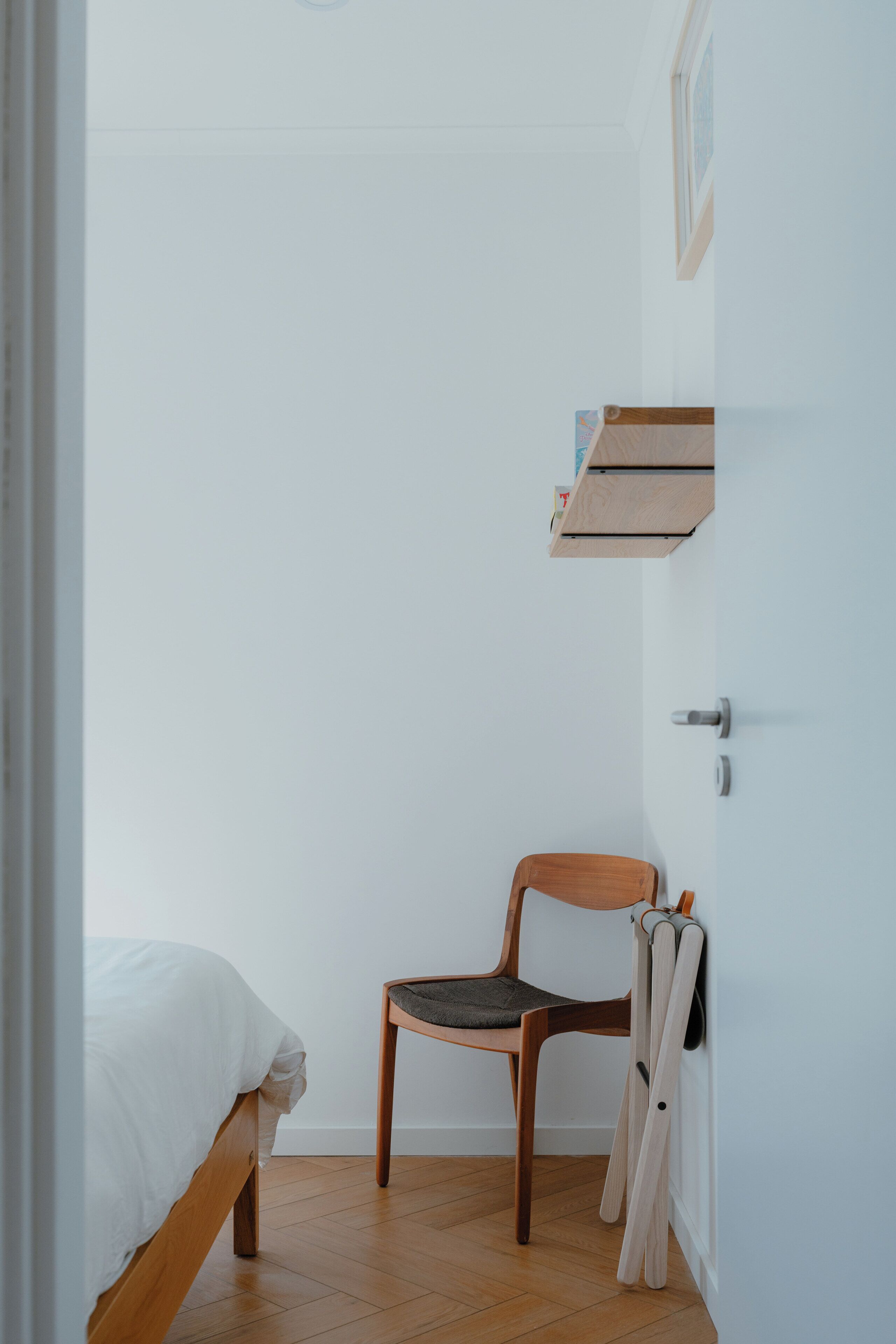 Een kamer met een bed, een houten stoel, een klein houten plankje en een deur.